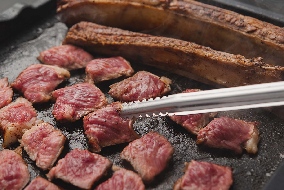 【下北沢店限定「自家燻製フンジェ王(ワン)カルビ」】
王(ワン)カルビと名付けた下北沢店の限定メニュー。韓国で「우대갈비(ウデカルビ)」と呼ばれる骨付き牛カルビを燻製し、香りと旨味を閉じ込めた自信作。鉄板でお好みの焼き加減に焼いていただき、まずはそのまま、お好みでニンニクやネギなどの薬味とともにサンチュで包んでお召し上がりいただきます